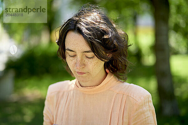 A young woman with freckles is captured in a sunny  natural setting  eyes closed and head slightly bowed  wearing a soft orange top.