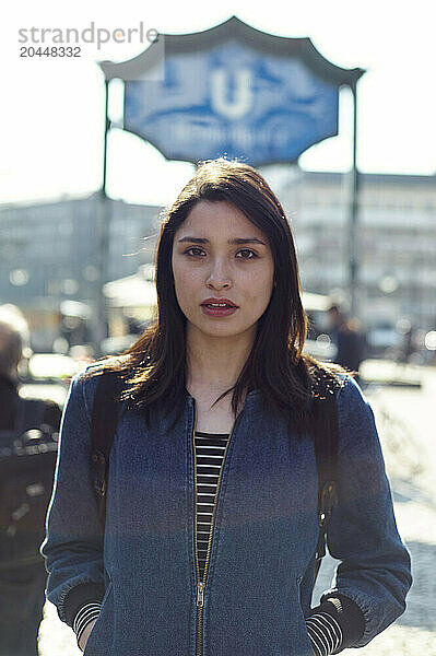 A young woman stands in front of a blurred city background with sunlight filtering through  with a signboard partially visible above her head.