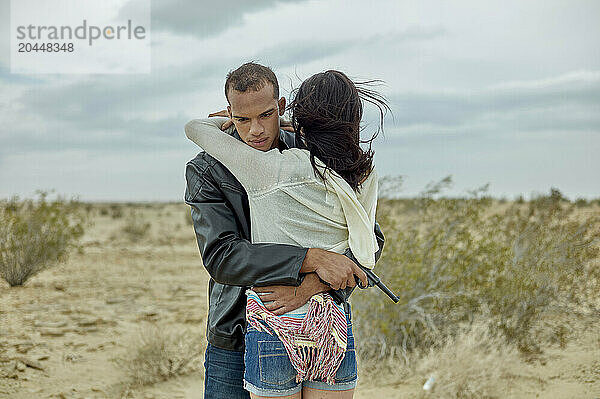 Two people embrace in a desert setting under an overcast sky  with sparse vegetation and a tranquil atmosphere.