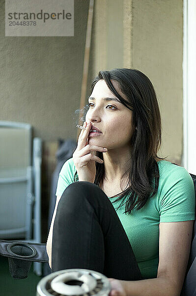 A woman is sitting indoors by a window with ample natural light  holding a cigarette and looking thoughtfully away from the camera.