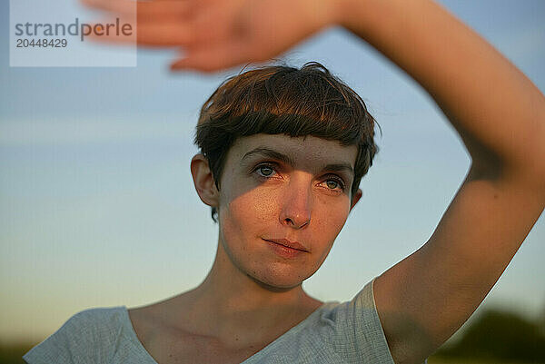 A young woman with short hair is photographed outdoors during twilight  with an out-of-focus hand gesturing near their face  creating an artistic effect.