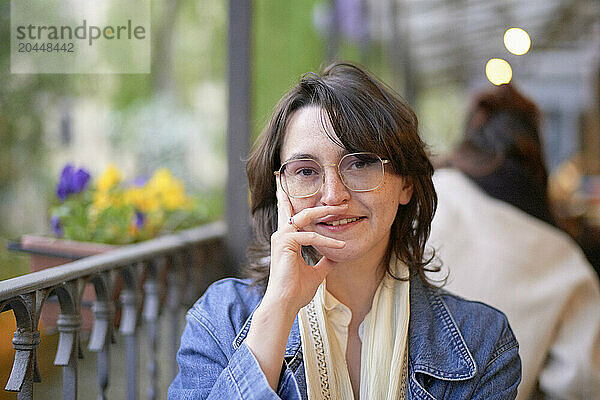 A smiling woman with glasses is sitting at an outdoor cafe  resting her chin on her hand.