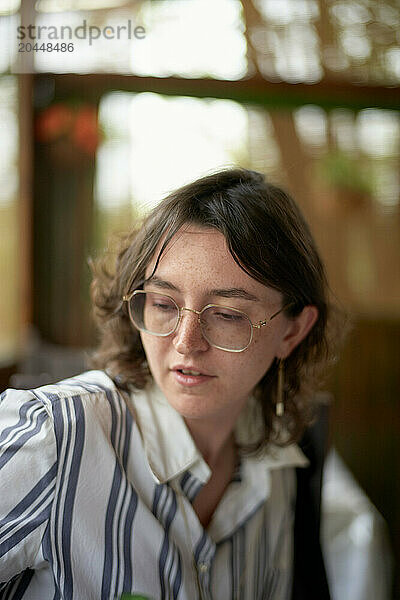 A young adult with glasses and short hair looks down thoughtfully  wearing a striped shirt and overalls against a blurred background.