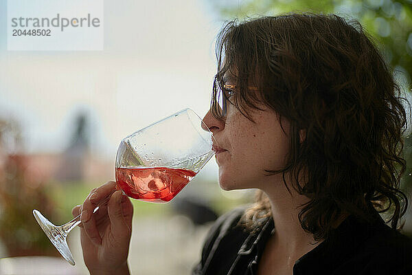 A woman is sipping wine from a large glass  with a blurred background suggesting an outdoor setting.