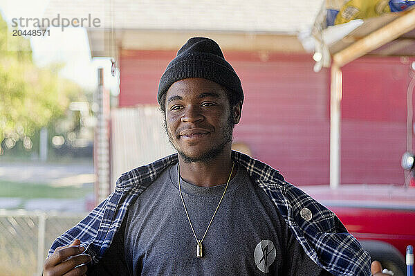 A smiling man wearing a beanie and plaid shirt stands in front of a house with a red door.