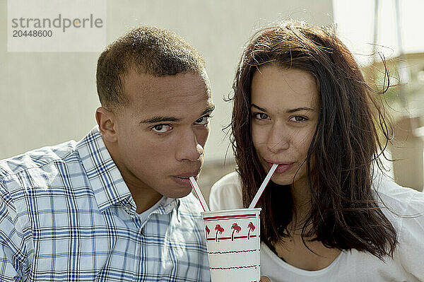 A young man and a woman are sharing a drink with two straws  looking towards the camera with a background that suggests they are outdoors in a slightly windy environment.