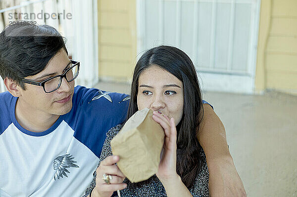 A woman is blowing into a brown paper bag  looking worried  while a man with glasses observes her closely.