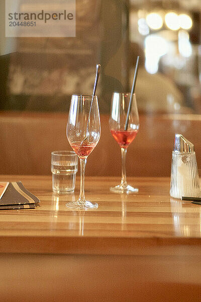 Two wine glasses  one partially filled with rose wine  sit on a wooden bar counter accompanied by a glass of water and a salt shaker  with a blurred background suggesting a cozy restaurant setting.