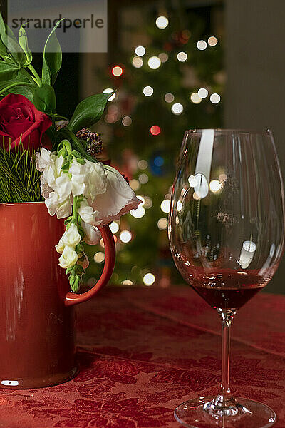 A festive table setting featuring a bouquet of flowers in a red vase and an empty wine glass  with a blurred Christmas tree adorned with lights in the background.