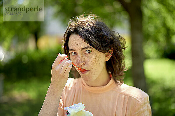 A person with freckles  wearing a peach-colored top  is eating a slice of orange  with a blurred green background.