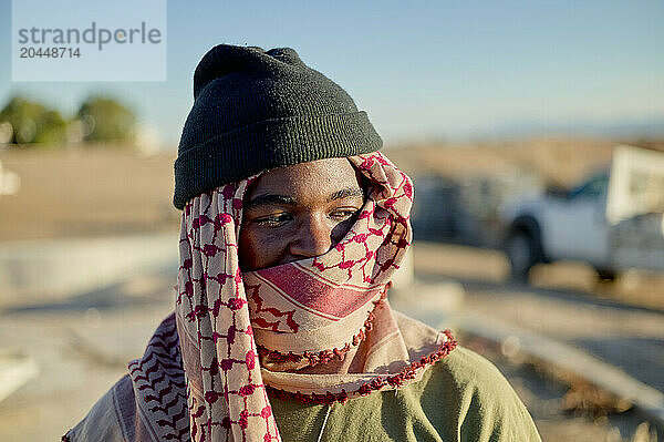 A person smiling  wrapped in a patterned scarf  wearing a beanie  and standing outdoors on a sunny day with vehicles and buildings in the blurred background.