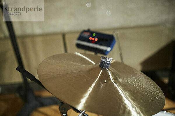 A close-up of a shiny cymbal as part of a drum set with a music amplifier lit by red LEDs in the soft focus background.