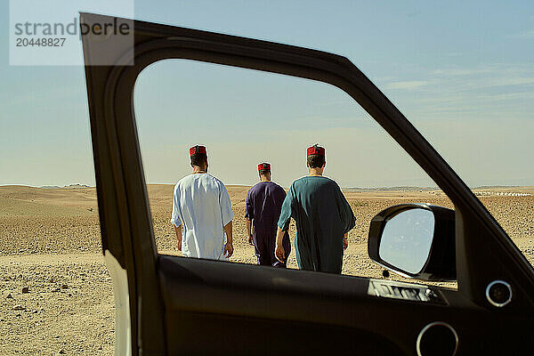 Three men in traditional attire walk through a desert landscape  viewed from a car's open door.