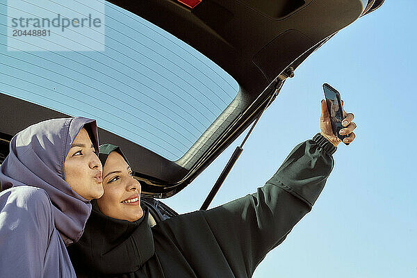 Two women in hijabs taking a selfie from the back of a car against a clear blue sky.