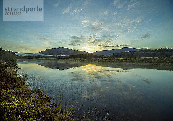 Sunrise over a tranquil lake with reflections of clouds and hills on the water surrounded by forested landscape.