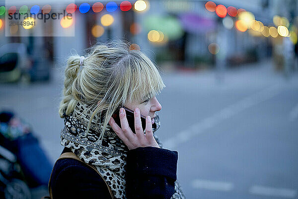 A woman talks on her phone while walking on a city street at dusk with colorful lights in the background.