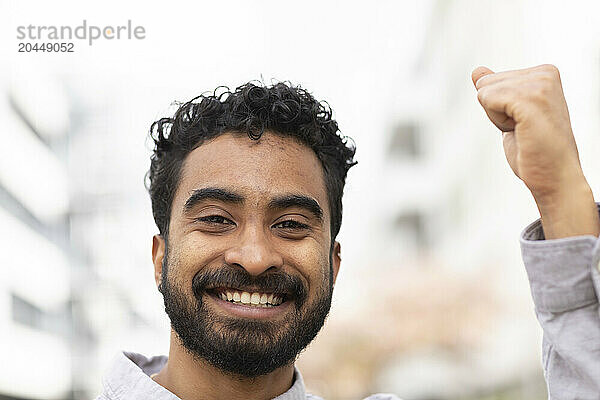 A smiling man with curly hair is raising his fist in a gesture of success or celebration against a blurred urban background.