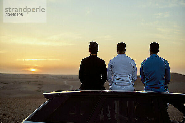 Three men sitting on a car watching sunset in the desert.