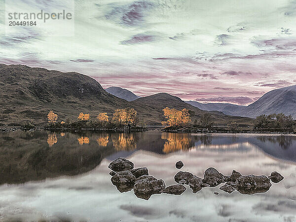 Tranquil sunset over a mirror-like lake with rocks in the foreground and mountains in the background under a colorful sky.