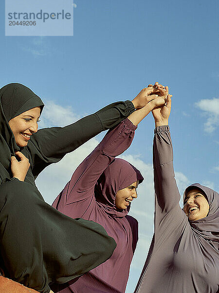 Three women in hijabs holding hands up together in solidarity under a blue sky.