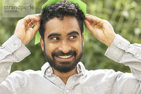 A smiling man with curly hair is putting on a green paper hat in a natural outdoor setting.