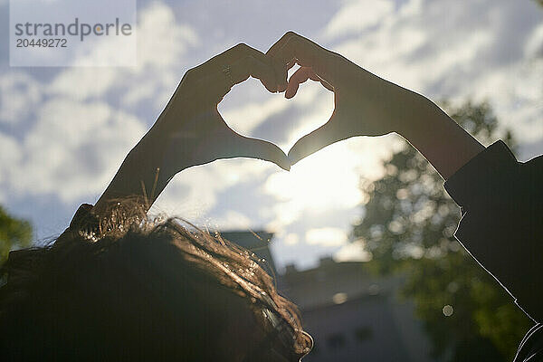 Silhouetted hands form a heart shape against a soft sunset sky.