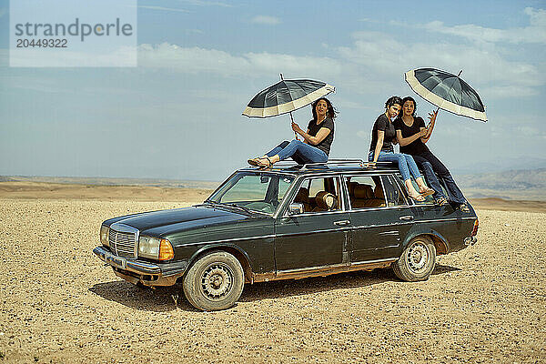 Group of friends having fun with umbrellas on a vintage car in a desert setting.