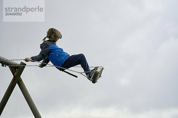 Child in a blue jacket enjoys swinging high against a cloudy sky backdrop.