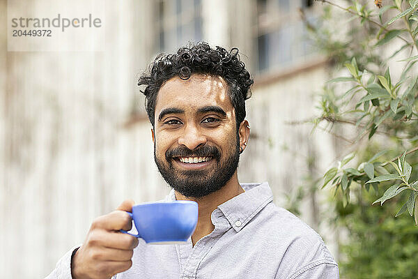 A smiling man with curly hair holds a blue cup towards the camera  standing in front of a blurred background