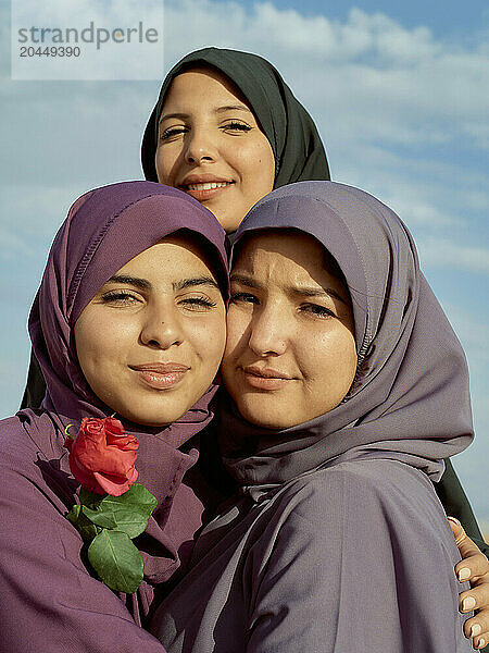 Three smiling women wearing hijabs embrace against a blue sky  one holding a red rose.