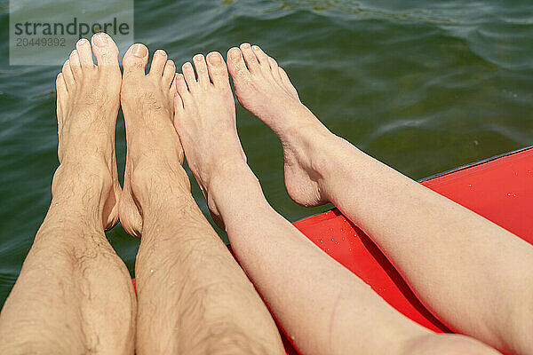 Two pairs of bare feet resting on the edge of a red boat on a water body