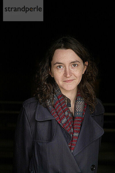 Smiling woman with curly hair wearing a gray coat and a patterned scarf against a dark background.