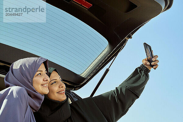 Two smiling women in headscarves taking a selfie with a smartphone by a car under a clear blue sky.