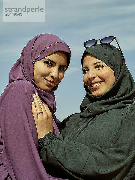 Two smiling women wearing hijabs posing under a clear blue sky.