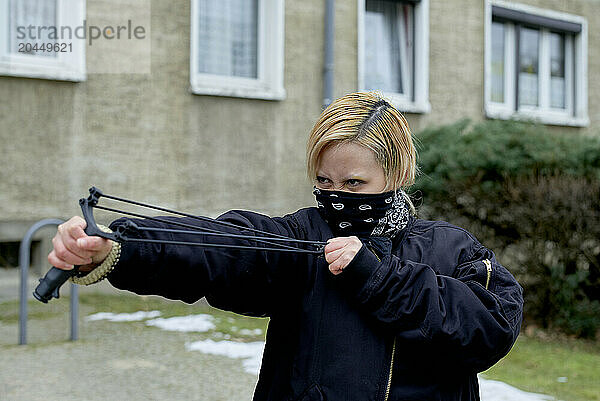 Woman in black with a bandana covering her face aiming a slingshot at the camera  with a building in the background.