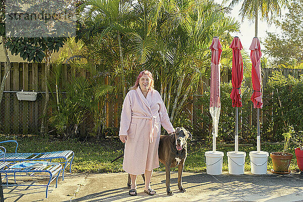 A smiling woman in a bathrobe stands beside a large dog in a sunny backyard with palm trees and folded patio umbrellas in the background.