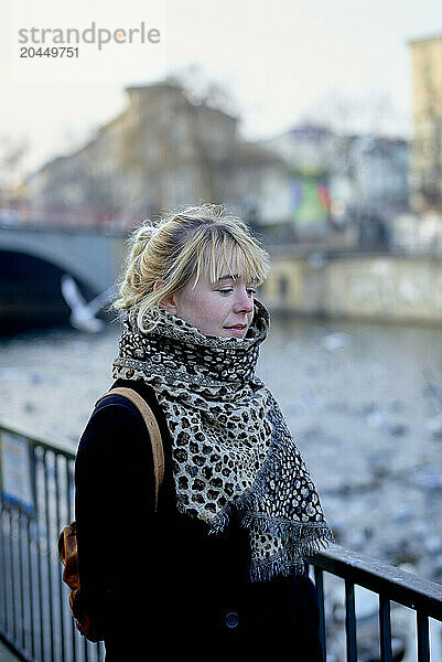 A young woman with a leopard print scarf and a backpack standing by a river railing with buildings and a bridge in the blurred background.