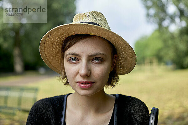 Woman with a straw hat and black top smiling in a park with green trees in the background.