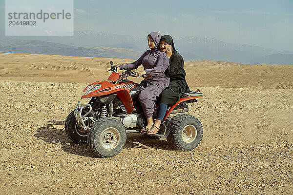 Two young girls smiling while sitting on a red quad bike in a desert landscape with mountains in the background.