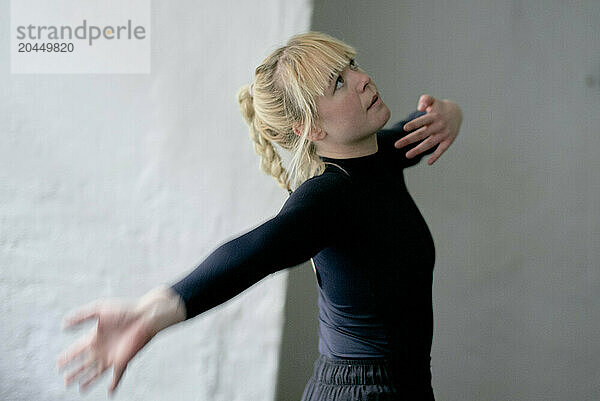 Young female dancer in black attire performing with expressive arm movements against a plain backdrop.