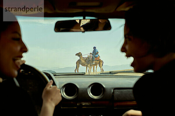 View from inside a car where two individuals in the front seats are conversing and laughing  with a camel rider seen through the windshield in a desert landscape.