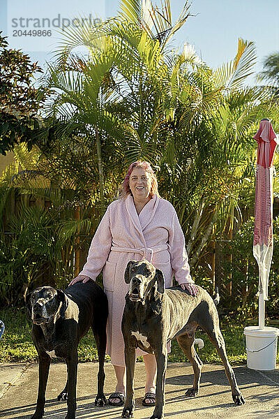 Smiling woman in bathrobe stands with two Great Danes on a sunny patio with palm trees in the background.