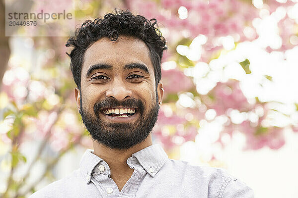 A smiling man with a beard and curly hair poses in front of a blooming pink flower tree.