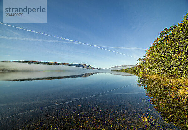 A serene lake with clear reflection under a blue sky  flanked by forest and low-lying fog. Loch Arkaig