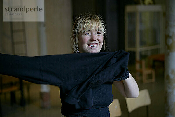 Smiling woman with blonde hair putting on a black shirt indoors.