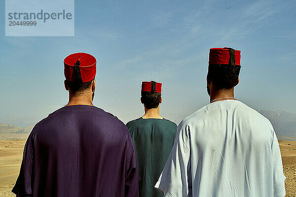 Three men in traditional attire stand side by side looking out over a desert landscape under a clear sky.