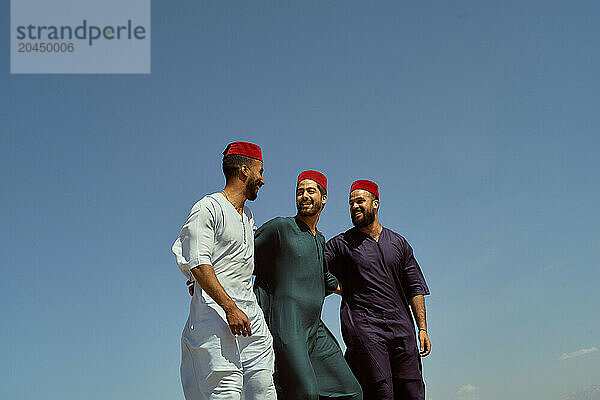 Three smiling men in traditional attire and red fezzes walk together under a clear blue sky.