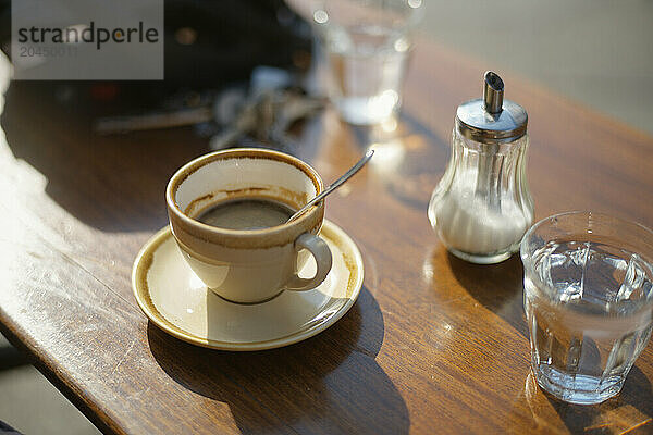 Morning coffee in a white cup with saucer  accompanied by a sugar dispenser and a glass of water  on a wooden tabletop bathed in sunlight.