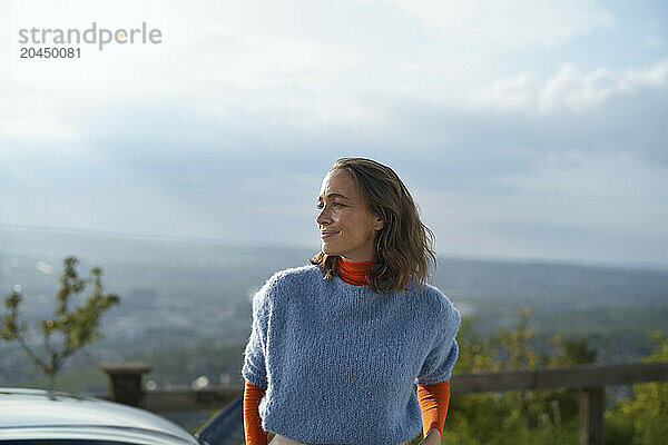 Smiling woman dressed in a blue sweater and orange shirt stands outdoors with a scenic overlook in the background.