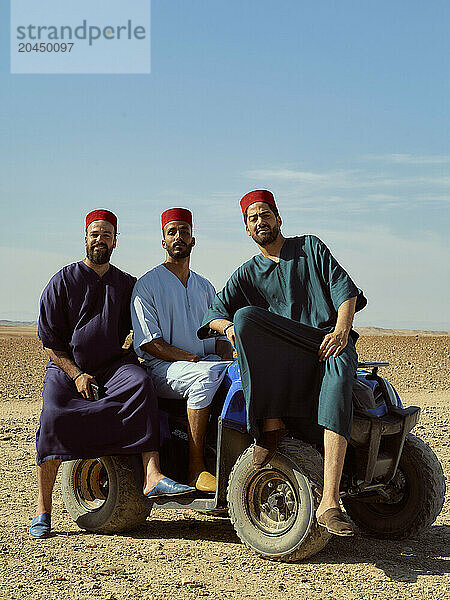 Three men wearing traditional fez hats pose confidently on a blue quad bike in a desert setting.